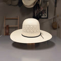 A cream-colored woven straw boater hat with a black band, displayed on a wooden stand on a gray table. The background is a workshop or shop interior with other hats hanging on the wall.