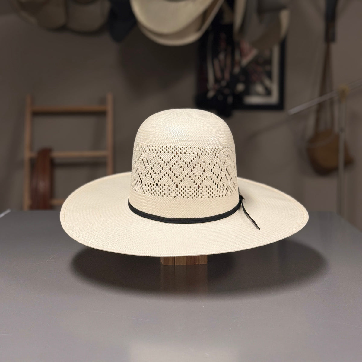 A cream-colored woven straw boater hat with a black band, displayed on a wooden stand on a gray table. The background is a workshop or shop interior with other hats hanging on the wall.