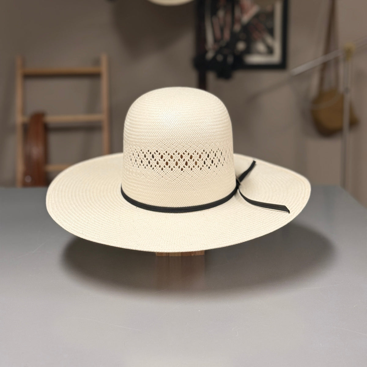 A cream-colored woven cowboy hat with a black ribbon band, displayed on a wooden stand on a gray table. The background shows a softly blurred interior with other hats hanging on a wall.