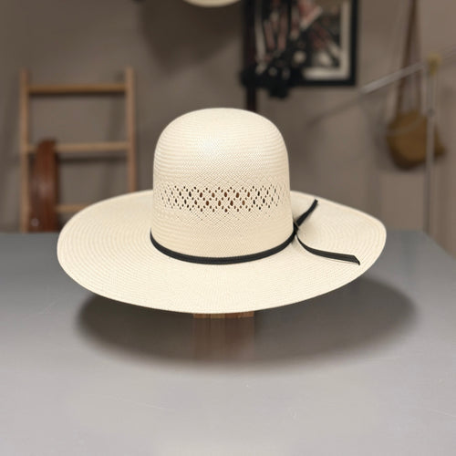 A cream-colored woven cowboy hat with a black ribbon band, displayed on a wooden stand on a gray table. The background shows a softly blurred interior with other hats hanging on a wall.