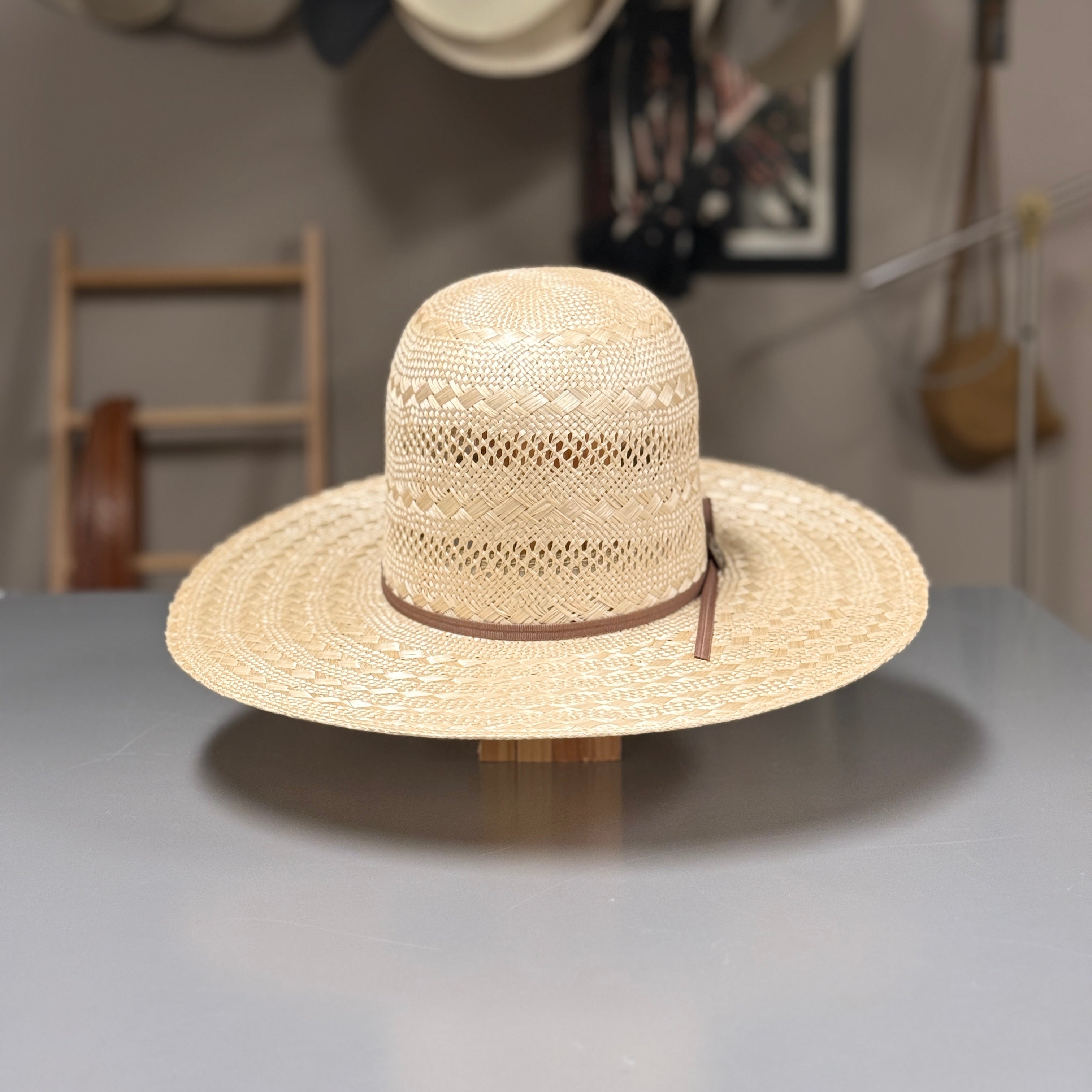A woven straw cowboy hat with a brown band sits on a display stand on a gray table, with a softly blurred shop interior in the background.