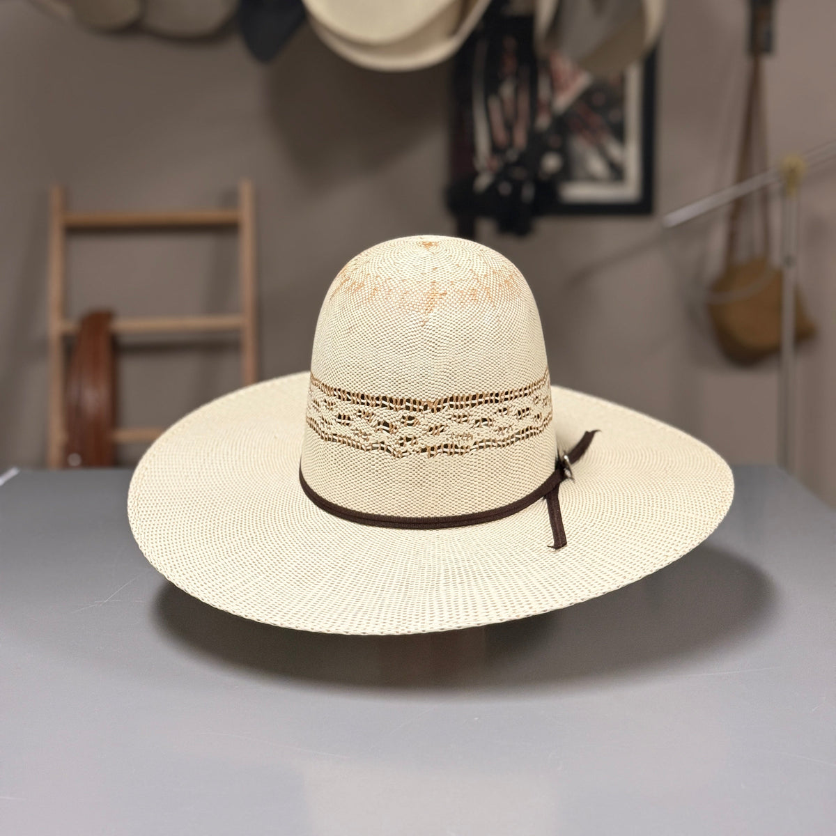 A straw cowboy hat with a wide brim, a decorative woven band, and a dark brown ribbon tie, displayed on a gray table in a hat shop.