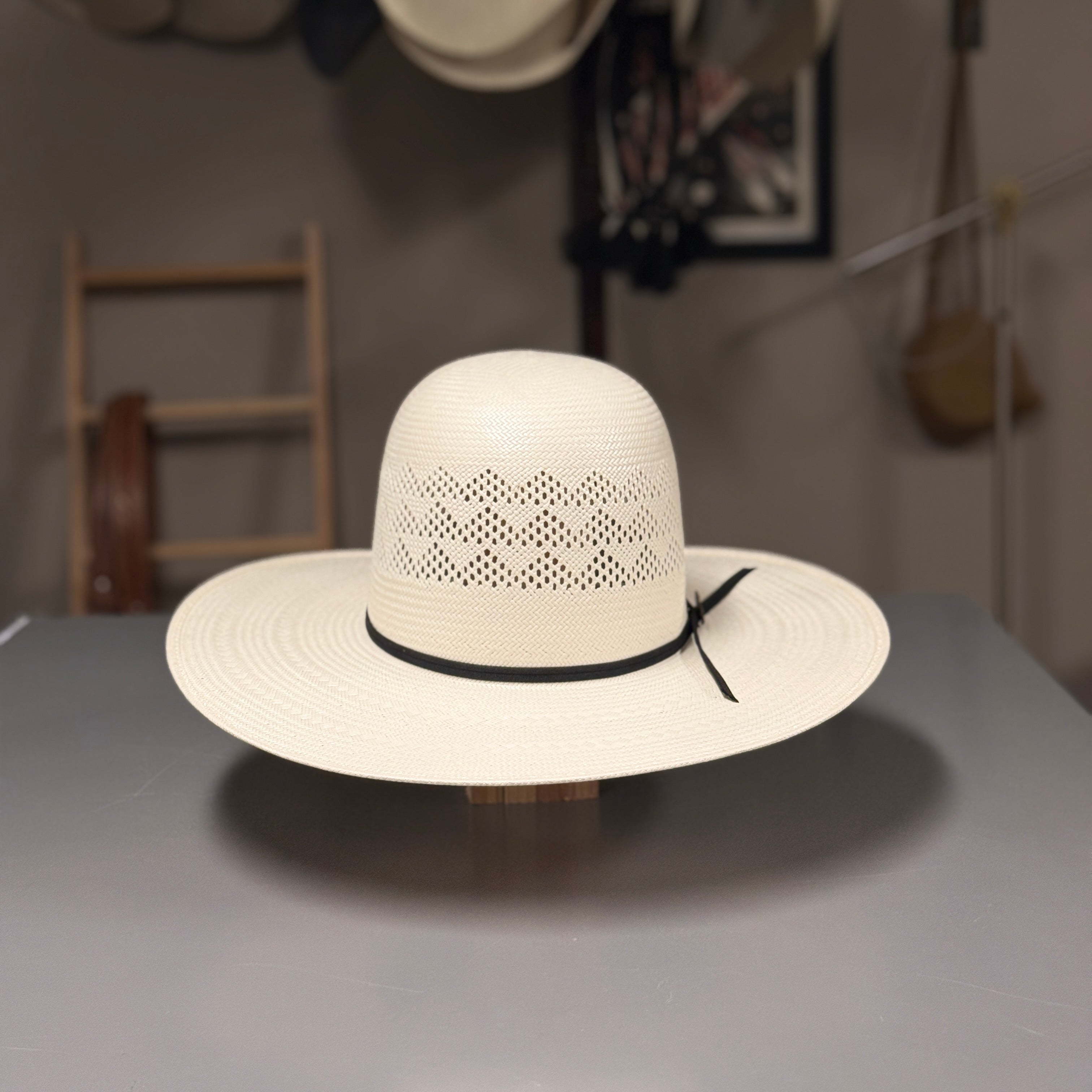 A cream-colored woven cowboy hat with a black band and bow, displayed on a stand on a gray table. The background shows a workshop setting with other hats hanging on a wall.