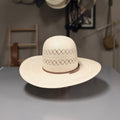 A cream-colored woven wide-brimmed cowboy hat with a decorative eyelet pattern on the crown and a brown band, displayed on a gray table in a shop setting with other hats hanging in the background.