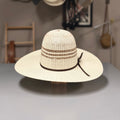 A cream-colored woven straw wide-brim hat with brown decorative bands and a matching ribbon tie, displayed on a stand on a gray table.